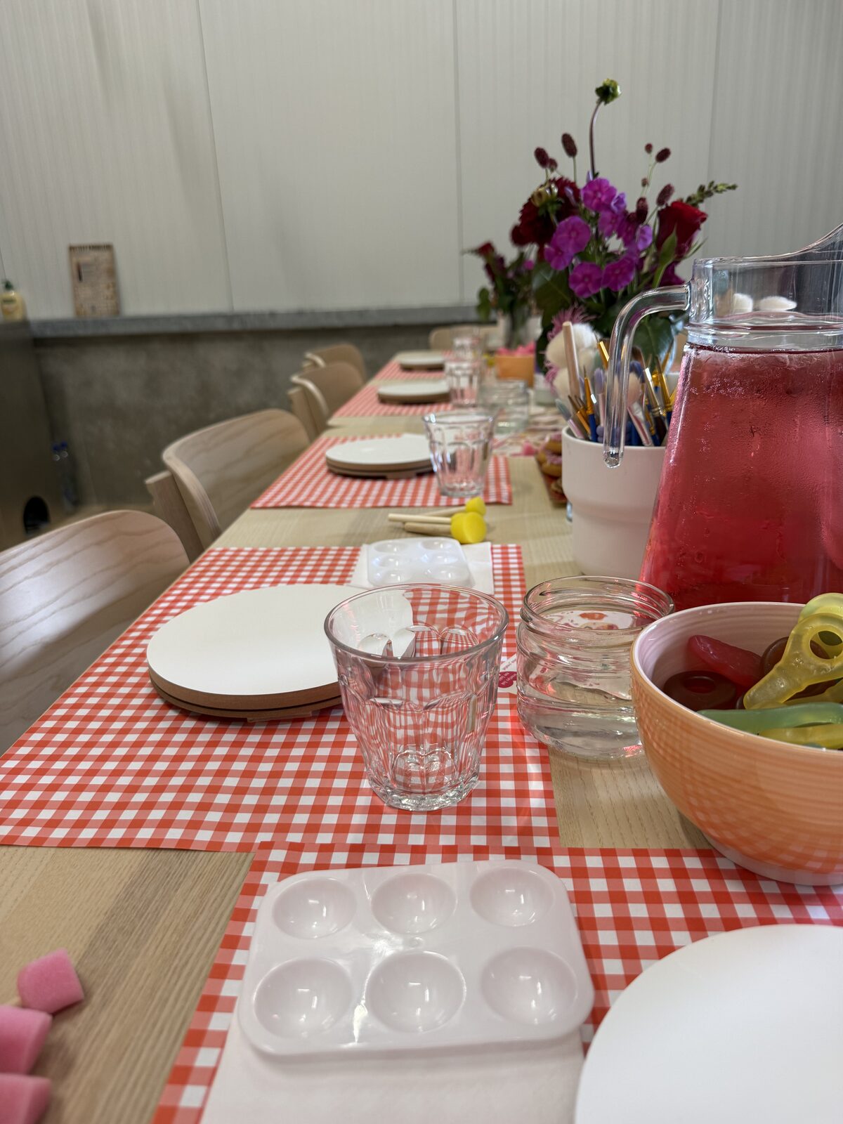 Gedekte werktafel met rood geruit tafelkleed, bloemen en drinken, klaar voor een workshop